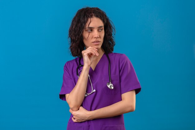 Mujer enfermera en uniforme médico y con estetoscopio sosteniendo la mano debajo de la barbilla con dudas sobre azul aislado