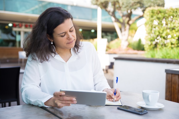 Mujer enfocada haciendo notas, trabajando y usando tableta en café