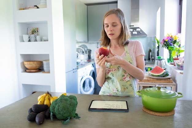 Foto gratuita mujer enfocada pensativa sosteniendo frutas mientras cocina en la cocina, usando tableta cerca de una cacerola y verduras frescas en el mostrador. vista frontal. cocinar en casa y concepto de alimentación saludable.