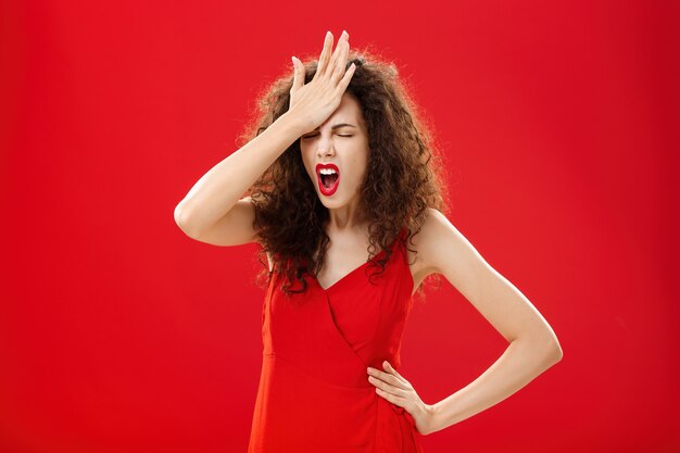 Mujer enojada con gente tonta. Mujer caucásica adulta olvidadiza irritada con el pelo rizado en un elegante vestido rojo que golpea la frente con la palma de la mano cerrando los ojos y jurando recordando algo importante.