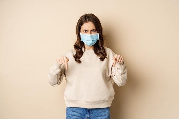 Mujer enojada con mascarilla médica, apuntando con el dedo hacia abajo y luciendo preocupada, parada en suéter sobre fondo beige