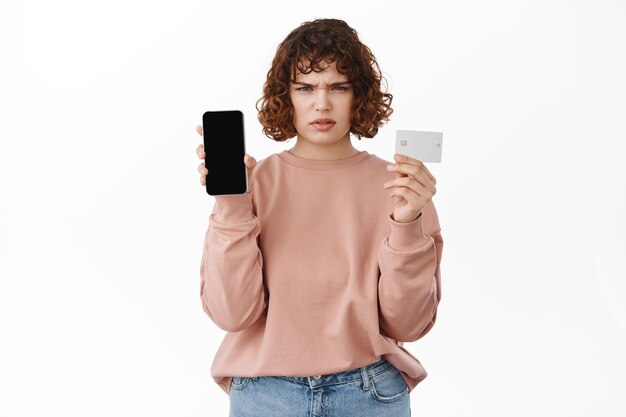 Mujer enojada mostrando tarjeta de crédito con pantalla de teléfono móvil, de pie con ropa informal.
