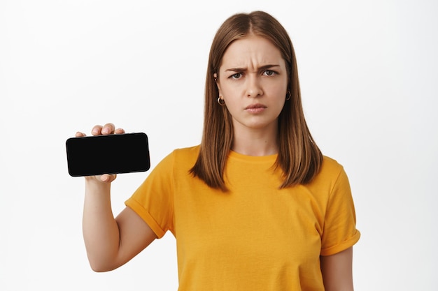 La mujer enojada muestra la pantalla horizontal del teléfono inteligente, frunce las cejas y parece molesta por el contenido del teléfono móvil, de pie disgustada contra la pared blanca