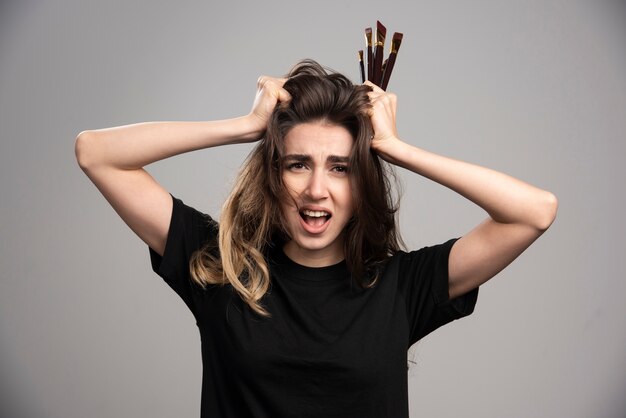 Mujer enojada sosteniendo cepillos tocando su cabello en la pared gris.