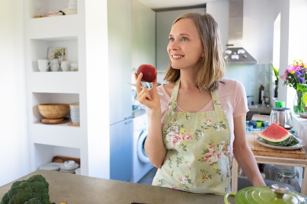 Mujer de ensueño alegre sosteniendo fruta, mirando a otro lado y sonriendo mientras cocina en su cocina. Copie el espacio. Cocinar en casa y concepto de alimentación saludable.