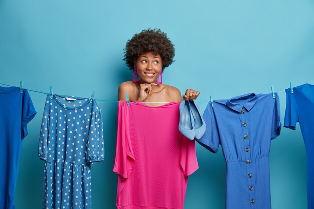 Una mujer de ensueño positiva con el pelo rizado se para desnuda y se cubre con un vestido colgando de una cuerda, sostiene zapatos azules, trata de encontrar un atuendo que le quede bien, se viste para ocasiones especiales. Gente, estilo, concepto de ropa.