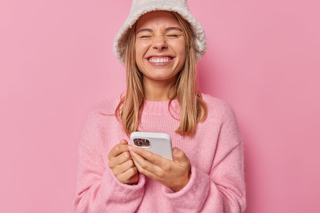 La mujer entrecierra los ojos y sonríe alegremente contenta de recibir el mensaje viste un jersey informal y un sombrero satisfecha con una aplicación increíble en rosa