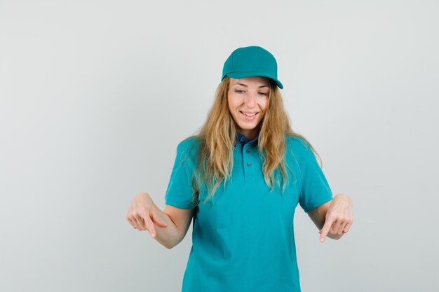 Mujer de entrega apuntando hacia abajo en camiseta, gorra y mirando alegre.