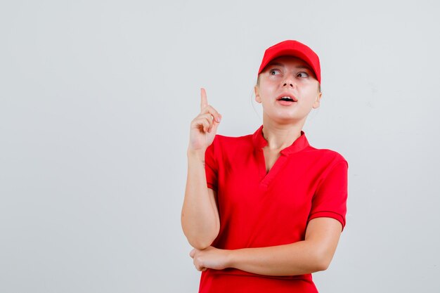 Mujer de entrega apuntando hacia arriba en camiseta roja y gorra y mirando curioso