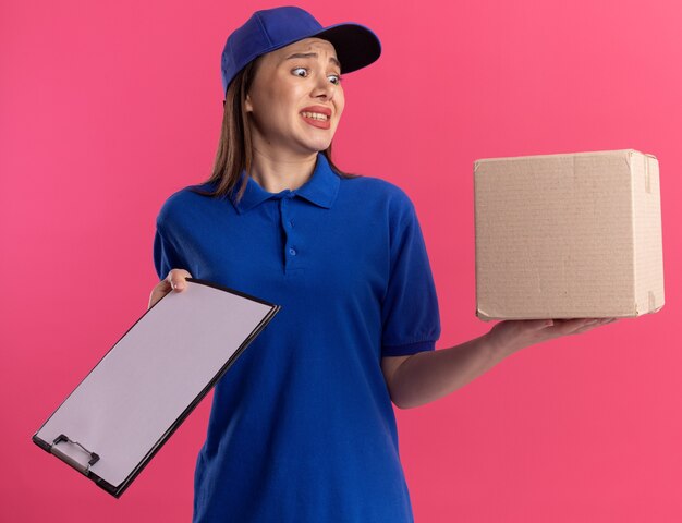 Mujer de entrega bonita ansiosa en uniforme sostiene el portapapeles y mira la caja de cartón
