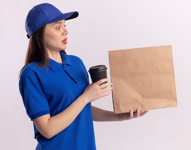 Mujer de entrega bonita ansiosa en uniforme sostiene la taza de papel y mira el paquete de papel en blanco