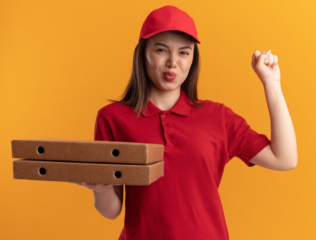 Mujer de entrega bonita complacida en uniforme mantiene el puño y sostiene cajas de pizza en naranja