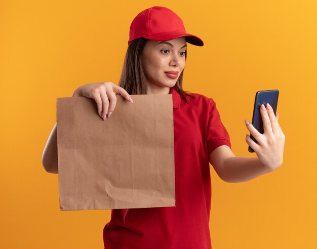 Mujer de entrega bonita complacida en uniforme sostiene el paquete de papel y mira el teléfono aislado en la pared naranja con espacio de copia