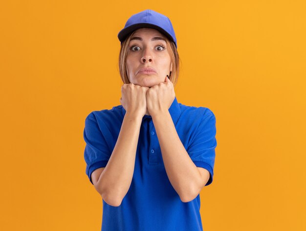 Mujer de entrega bonita joven ansiosa en uniforme pone los puños en la barbilla y mira al frente aislado en la pared naranja