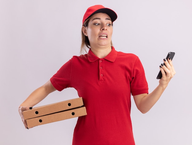 Mujer de entrega bonita joven ansiosa en uniforme sostiene cajas de pizza y mira teléfono aislado