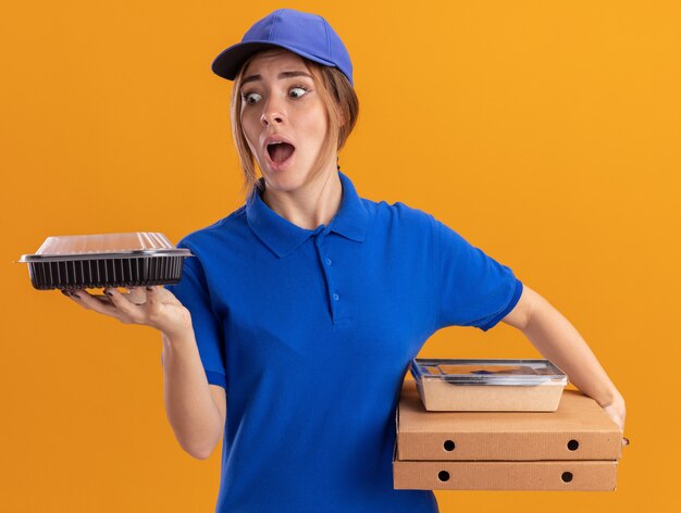 Mujer de entrega bonita joven ansiosa en uniforme sostiene paquetes de alimentos de papel y contenedores en cajas de pizza aisladas en la pared naranja