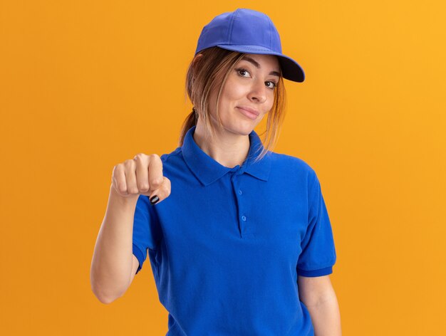 Mujer de entrega bonita joven complacida en uniforme mantiene el puño y mira al frente aislado en la pared naranja