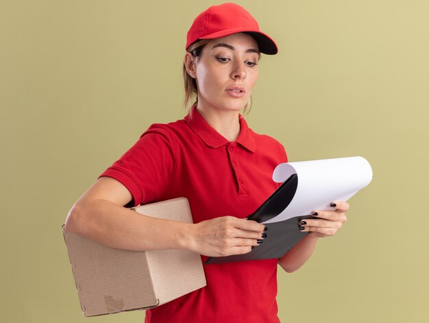 Mujer de entrega bonita joven confiada en uniforme sostiene la caja de cartón y mira el portapapeles aislado en la pared verde oliva