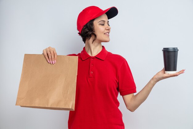 Mujer de entrega bonita joven contenta que sostiene el empaquetado de papel de los alimentos y que mira la taza para llevar