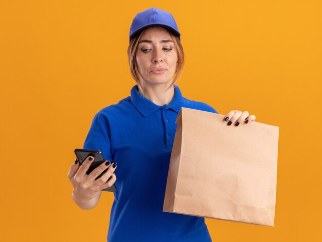 Mujer de entrega bonita joven decepcionada en uniforme sostiene el paquete de papel y mira el teléfono aislado en la pared naranja