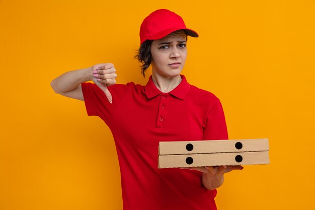 Mujer de entrega bonita joven disgustada sosteniendo cajas de pizza y pulgar hacia abajo