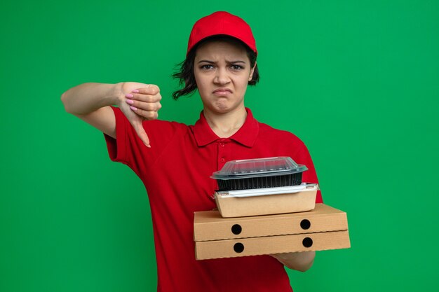 Mujer de entrega bonita joven disgustada sosteniendo contenedores de alimentos con envases en cajas de pizza y pulgar hacia abajo