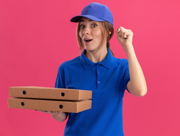 Mujer de entrega bonita joven emocionada en uniforme levanta el puño y sostiene cajas de pizza aisladas en la pared rosa