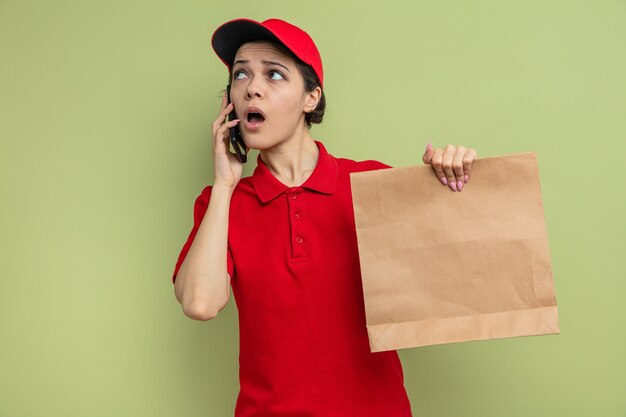Mujer de entrega bonita joven sorprendida hablando por teléfono y sosteniendo envases de papel para alimentos mirando a un lado