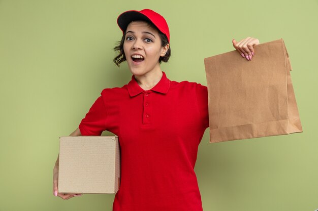 Foto gratuita mujer de entrega bonita joven sorprendida que sostiene el empaquetado de papel de los alimentos y la caja de cartón