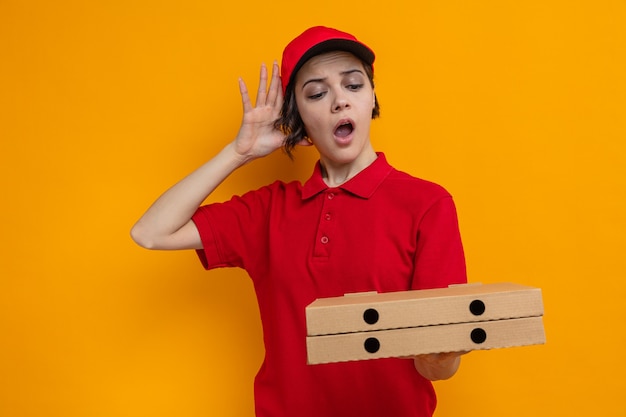 Mujer de entrega bonita joven sorprendida sosteniendo y mirando cajas de pizza