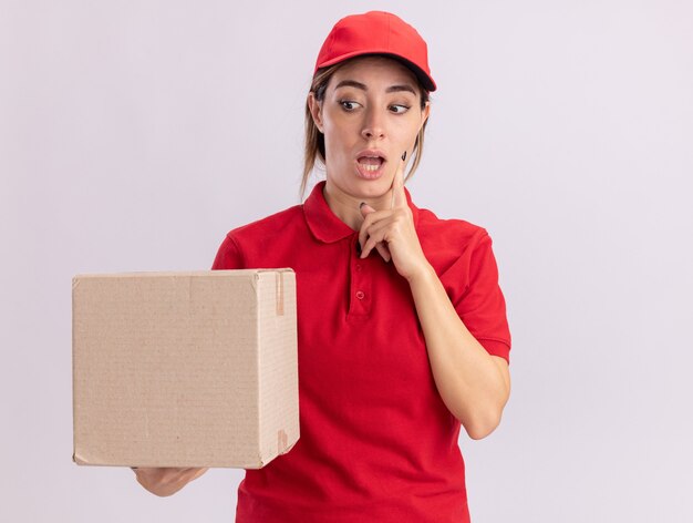 Mujer de entrega bonita joven sorprendida en uniforme pone el dedo en la cara sosteniendo y mirando la caja de cartón aislada en la pared blanca