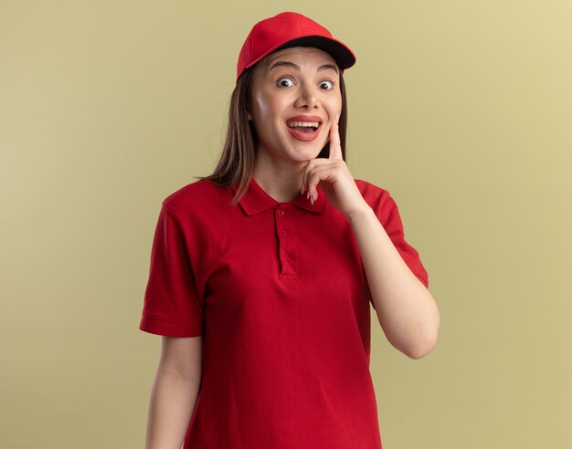 Mujer de entrega bonita sorprendida en uniforme pone el dedo en la cara y mira a la cámara