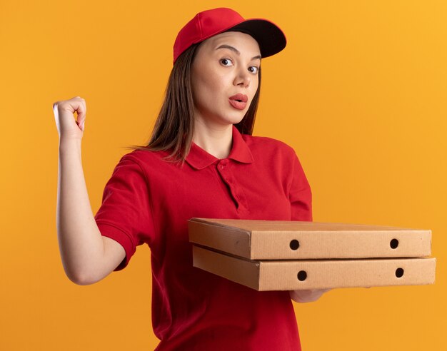 Mujer de entrega bonita sorprendida en uniforme sostiene cajas de pizza y apunta hacia atrás