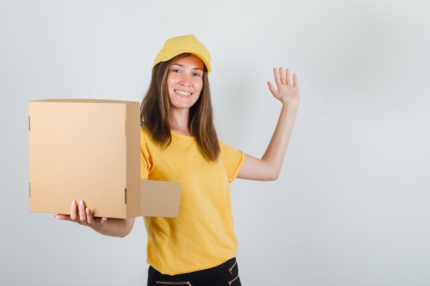 Mujer de entrega con caja abierta con signo de mano en camiseta, pantalón, gorra y mirando contento