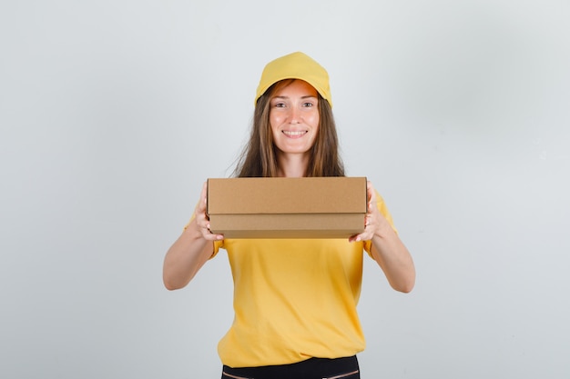 Mujer de entrega con caja de cartón en camiseta amarilla, pantalones y gorra y mirando alegre