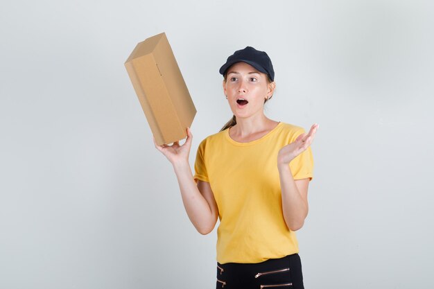 Mujer de entrega con caja de cartón en camiseta, pantalón y gorra y mirando sorprendido