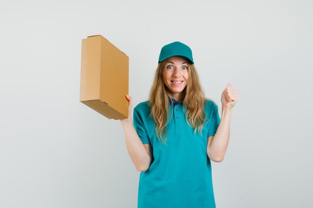 Mujer de entrega con caja de cartón con gesto de ganador en camiseta, gorra y mirando feliz