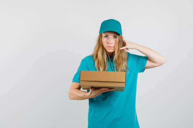 Mujer de entrega con caja de cartón con gesto de teléfono en camiseta, gorra