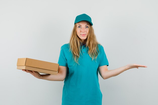 Mujer de entrega con caja de cartón con palma extendida en camiseta, gorra y mirando feliz.