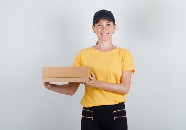 Mujer de entrega con caja de cartón y sonriendo en camiseta, pantalón y gorra