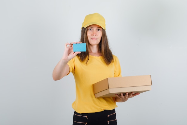 Mujer de entrega con caja de cartón y tarjeta azul en camiseta, pantalón y gorra y mirando contento