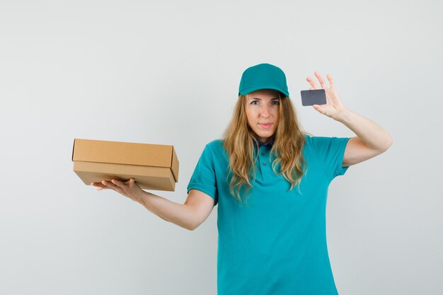 Mujer de entrega con caja de cartón y tarjeta en camiseta, gorra y mirando alegre.