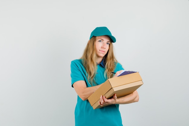 Mujer de entrega en camiseta, gorra escribiendo en portapapeles en caja de cartón