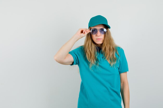 Mujer de entrega en camiseta, gorra mirando a cámara a través de gafas y mirando enfocado