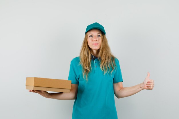 Mujer de entrega en camiseta, gorra sosteniendo una caja de cartón con el pulgar hacia arriba y mirando alegre