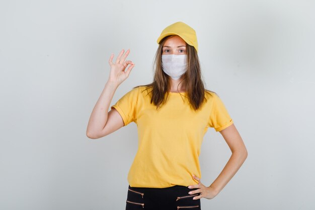 Mujer de entrega en camiseta, pantalón, gorra, máscara que muestra un gesto bien y parece contento