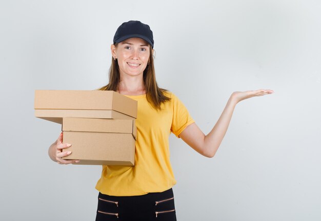 Mujer de entrega en camiseta, pantalón, gorra sosteniendo cajas de cartón y sonriendo