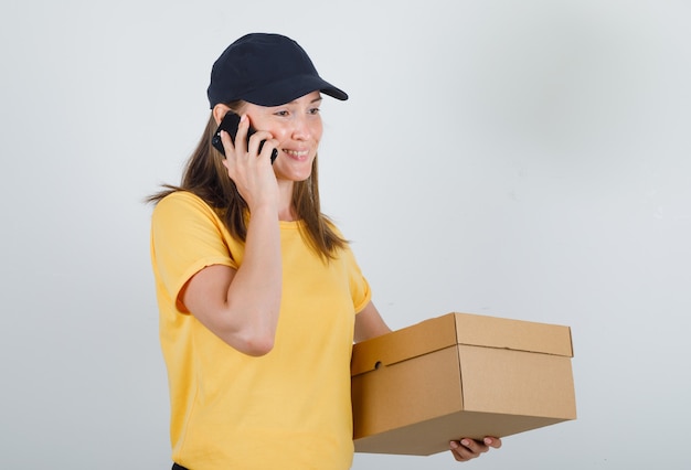 Mujer de entrega hablando por teléfono inteligente con caja de cartón en camiseta, pantalón y gorra