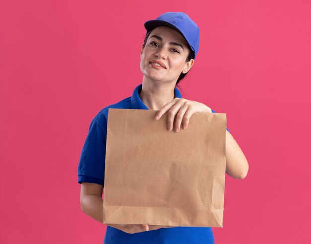 Mujer de entrega joven complacida en uniforme y gorra sosteniendo el paquete de papel aislado en la pared rosa con espacio de copia