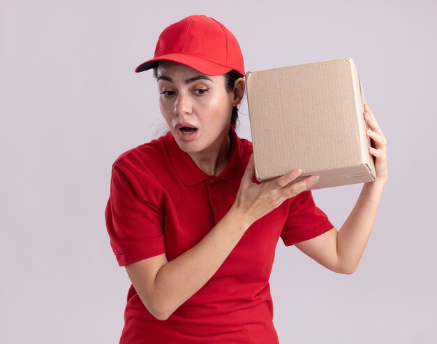 Mujer de entrega joven curiosa en uniforme y gorra sosteniendo una caja de cartón cerca de la oreja mirando hacia abajo escuchando